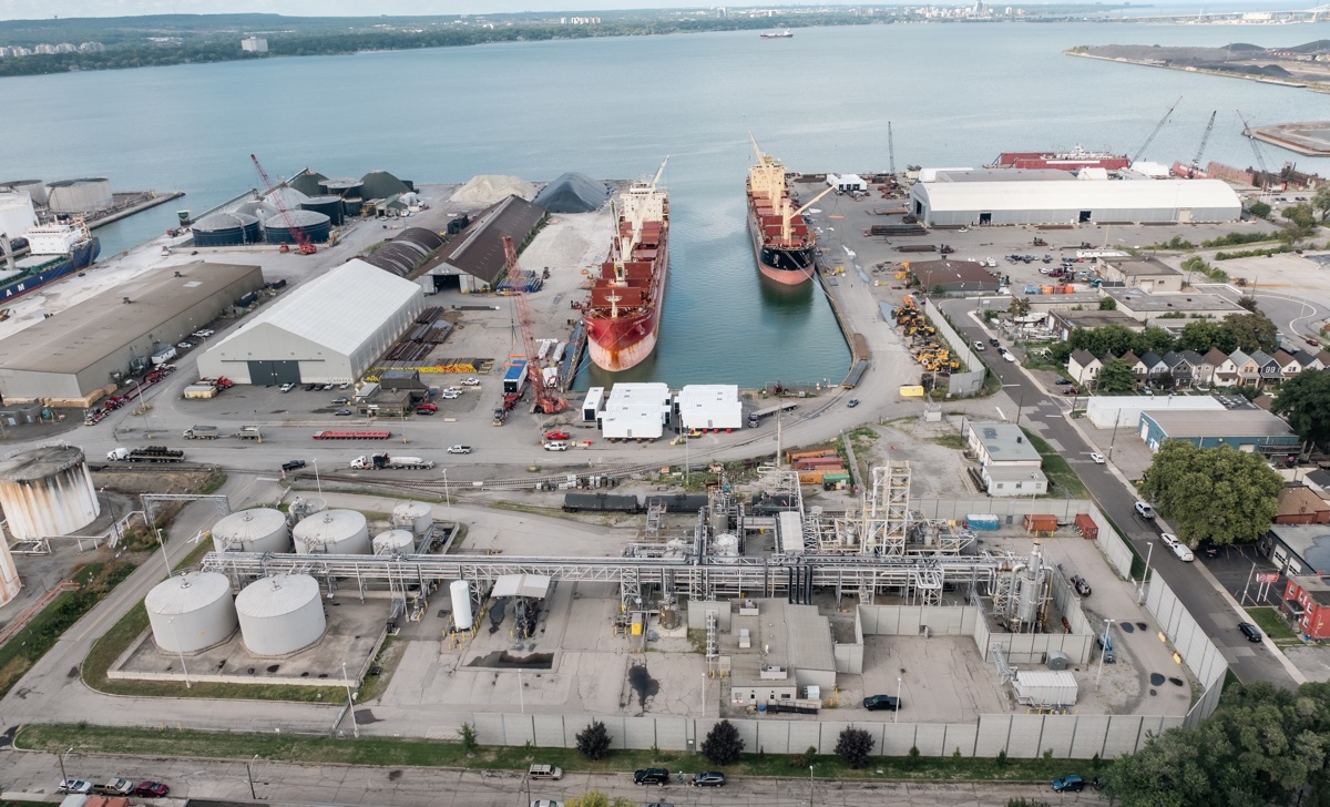 Aerial overview of the Biidaaban biodiesel facility in Hamilton, with storage tanks and process equipment in the foreground, rail infrastructure, port berths, ships, and Hamilton Harbour beyond.